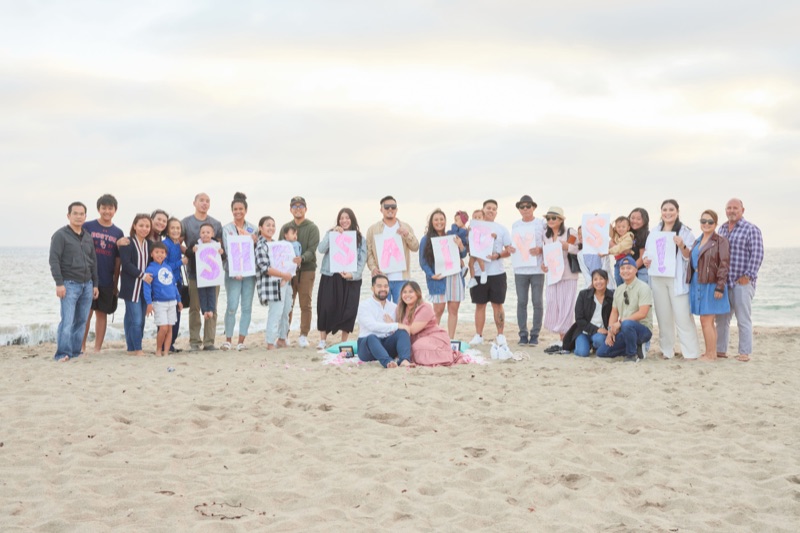 Both families on the beach holding SHE SAID YES signs behind Debra and Vinh